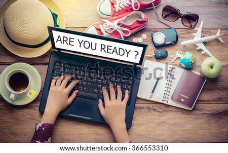 Hands on the keyboard of a notebook's screen displays, word "ARE YOU READY ?" and travel accessories are placed on a wooden table. The concept plan tour