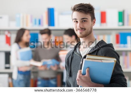 Confident handsome student holding books and smiling at camera, library bookshelves on background, learning and education concept Royalty-Free Stock Photo #366568778