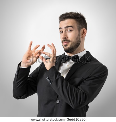 Young proud groom holding and showing engagement ring looking at camera. Portrait over gray studio background. 