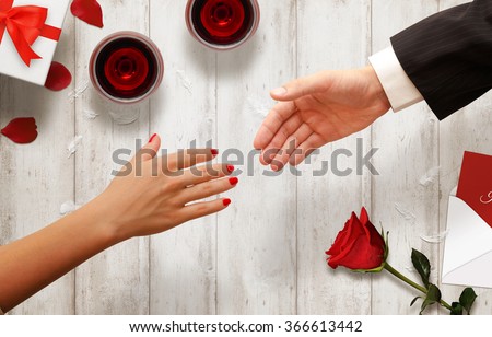 Couple gives each other a hand. Background with glasses of wine, gift, rose, envelope on wooden table.