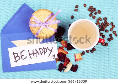 Cup of coffee with milk on the table. Morning coffee, cookies, rose petals, coffee beans and blue envelope with text "Be happy"
