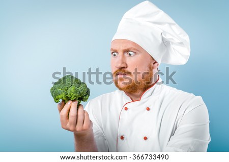 Funny picture of young male chef in white uniform. Head-cook looking at broccoli with bulging eyes. Standing against grey background