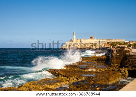 Cuba. Island. Ocean. Sea. Green and brown stones. High lighthouse. Waves breaking on the shore. White water spray. Blue sky. Nature