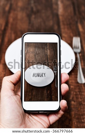 closeup of a young caucasian man taking a picture with his smartphone of a plate with the word hungry written in it, placed on a wooden table