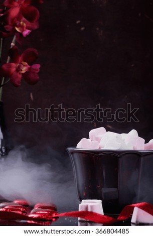 Heart shaped St Valentine's candies and marshmallows in grey ceramic bowl with smoke and purple orchid over the black background