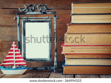 stack of old books next to decorative sailing boat and blank frame wooden table. vintage filtered image