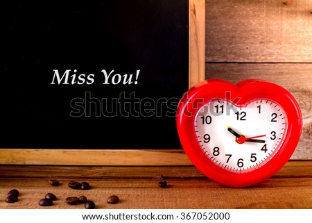 Red alarm clock with heart shape and blakc board with message Miss you. 