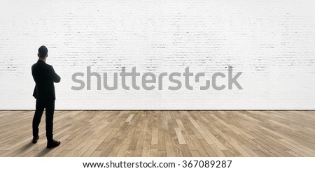 Businessman stands opposite bricks wall in gallery interior with wooden floor.  Royalty-Free Stock Photo #367089287