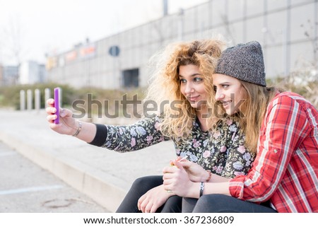 Two young curly and straight blonde hair caucasian woman sitting on a sidewalk, using a smartphone, taking a selfie - technology, social network, communication concept