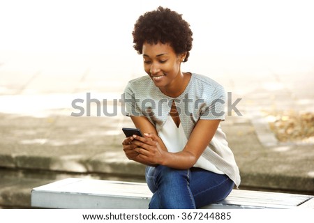Portrait of happy young african woman sitting outside on bench reading a text message on her mobile phone Royalty-Free Stock Photo #367244885