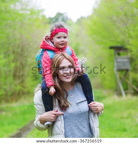 Beautiful mother carrying little girl on shoulders. Happy family of two, young smiling woman and her cute toddler daughter enjoying beautiful day in the forest