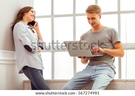 Happy couple uses laptop and talking on the phone while standing near the windowsills at home