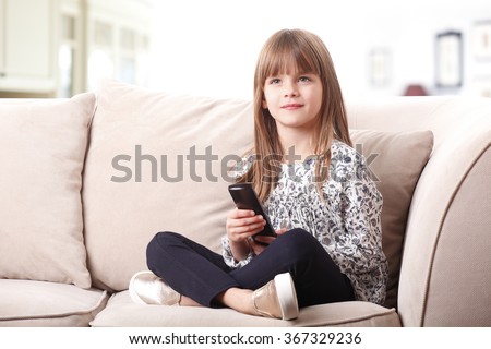 Portrait of adorable little girl sitting on couch and holding in her hand a remote control while watching cartoon at tv.