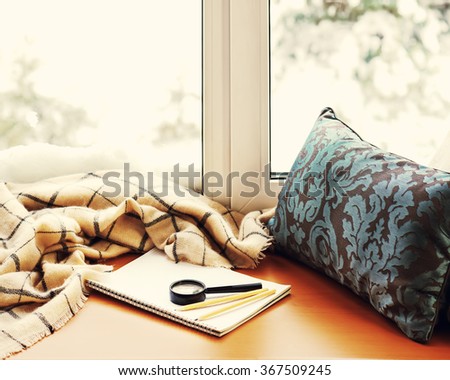Open notepad, magnifier glass, pillow, pencils and beige warm plaid located on stylized wooden windowsill. Winter concept of comfort and relaxation. Photo with retro filter effect.