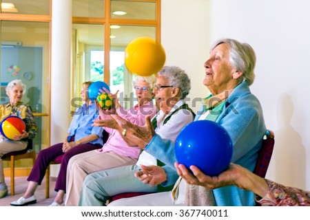 Group of happy senior ladies doing coordination exercises in a seniors gym sitting in chairs throwing and catching brightly colored balls Royalty-Free Stock Photo #367740011