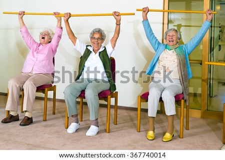 Three happy elderly ladies doing exercises in a seniors gym sitting in chairs raising wooden poles above their heads while smiling and laughing Royalty-Free Stock Photo #367740014