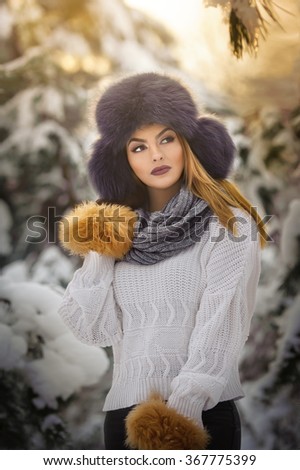 Beautiful woman in white pullover with over-sized fur cap enjoying the winter scenery in forest. Blonde girl posing under snow-covered trees branches.Attractive young female in bright cold day, makeup
