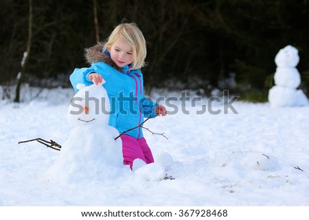 Cute toddler girl in warm snowsuit playing with a snow. Little kid play having fun outdoors building snowman in the forest. 