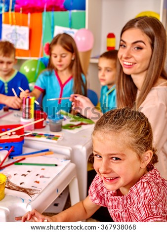 Large group of kids with female teacher painting on paper at table in kindergarten . 