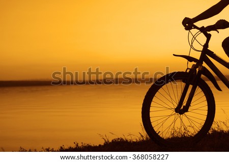 silhouette of woman's hands holding wheel of bicycle at sunset cloudy sky background and reflection in water. No face. Unrecognizable person. Empty copy space for inscription.