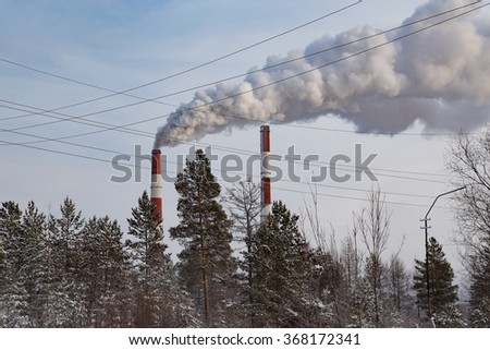 Two smokestacks with thick smoke, trees and wires in the foreground