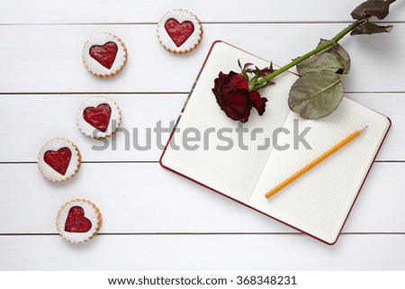 Heart shaped cookies with empty notebook, pencil and rose flower on white wooden background for Valentines day celebration.