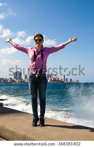 young woman with red hair in sunglasses, pink shirt, jeans, photographer. camera. hands up. smile. tourist. stand on gray stone. Beach Ocean Sea. blue water. splashes of white. wave. sky. cloud. City