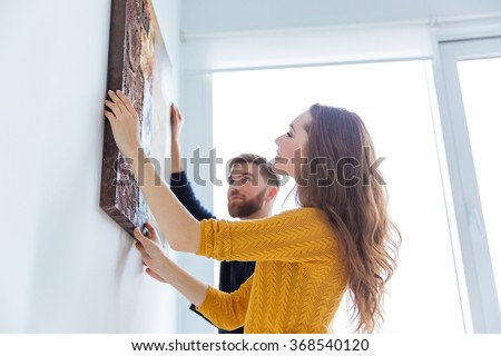 Happy couple hanging picture on the wall at home Royalty-Free Stock Photo #368540120