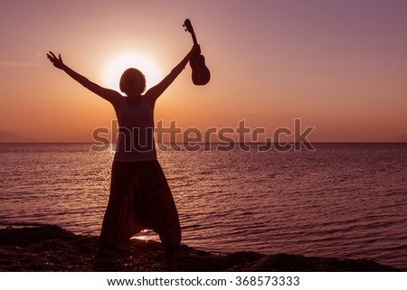 Silhouette of young girl standing on the beach with hands up, with ukulele, at the sunset against the sun