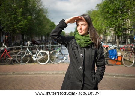 Beautiful young woman admiring the beauty of Amsterdam on a bridge protect their eyes from sun with hand dressed in black coat and green scarf