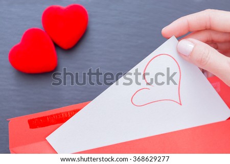 Woman getting from envelope paper sheet with pictured heart. Two red decorative hearts on grey background. Unrecognizable