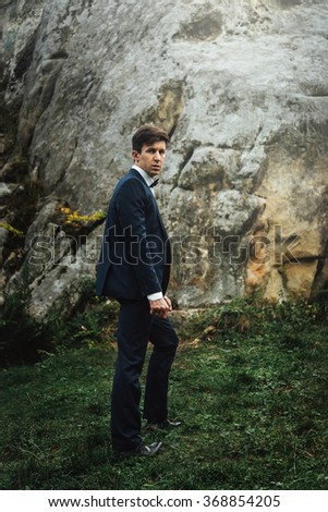 stylish elegant happy groom in fashionable suit posing on background of rocks in mountains