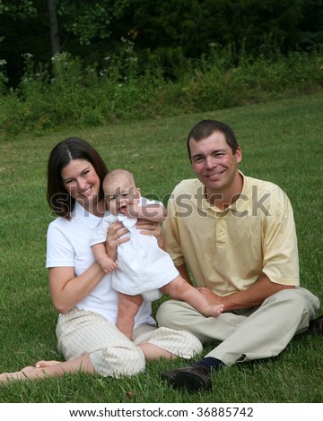 Family with a  4 month old baby poses in the park.  Here they are sitting on the grass.