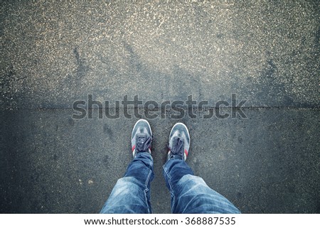 Alone man standing on textured grunge asphalt city street, point of view perspective. Pedestrian with blue shoes and jeans pants standing alone on grunge pavement. Royalty-Free Stock Photo #368887535