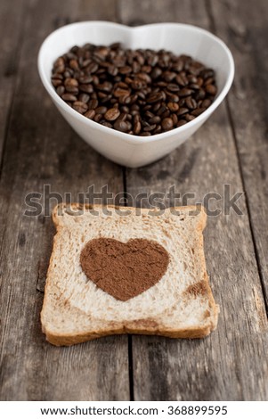 Bread slice with heart shape and coffee beans in white bowl on wooden background.