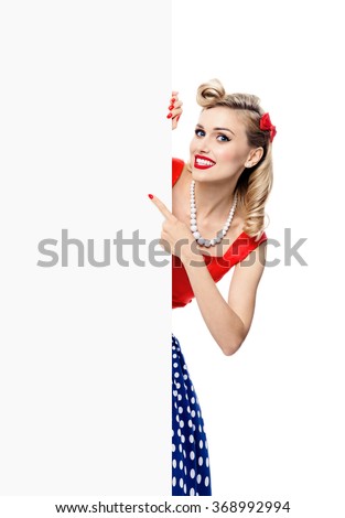 Portrait of smiling blond woman in pin-up style dress, showing blank signboard with copyspace area for slogan or text. Caucasian blond model posing in retro fashion and vintage concept studio shoot.