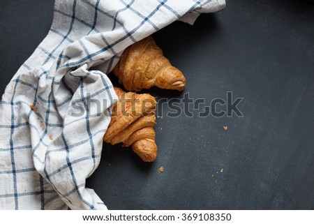 Two fresh croissant lying on a blackboard. Also, there is twisted linen towel. Croissants are delicious and appetizing. Towel is white with blue pattern.