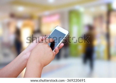 hand hold and touch screen smart phone, on blurred photo of department store shopping mall center and people background