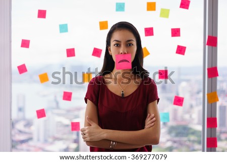 Mixed race woman working in modern office with reminders on skyscraper window. The girl feels stressed, holds a sticking note with sad emoticon on mouth.