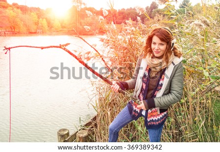 Young model woman with earmuffs playing on lake shore - Teenager girl on winter vacation having fun fishing in a green water river - Concept of alternative leisure activities - Gaussian blur filter