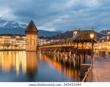 Kapellbrucke Chapel Bridge and Water Tower under Dramatic Sky Illuminated Building Reflection from Reuss River Sceneic Night Panorama Cityscape Skyline. Covered bridge in Lucerne, Switzerland