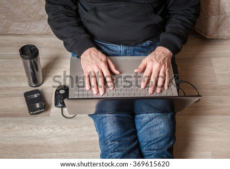 man sits on a wooden floor with a laptop on hir feet . the phone  and a cup tumbler with coffee