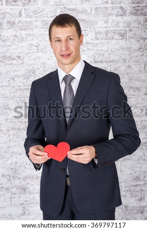 A smiling man in a suit with a red heart on a hand on a white brick wall background. The young man, a guy with a red heart. Valentine's Day, give love.