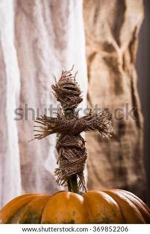 Closeup photo of handmade cross shaped Halloween sackcloth witching doll put on stem of round orange segmented cucurbita on white and brown fabric background, vertical picture