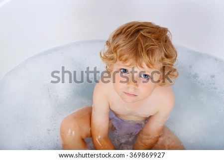 Adorable blond toddler boy playing and swimming by taking bath in bathtub. angry kid looking at the camera. Carefree childhood.