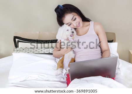 Picture of attractive young girl working on the laptop while hugging her puppy in the bedroom