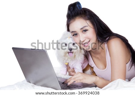 Picture of pretty young woman smiling at the camera while using notebook on bed with her dog