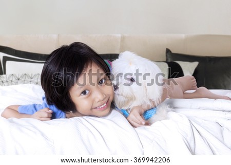 Picture of happy little girl smiling at the camera while lying on the bed and hugs a maltese dog