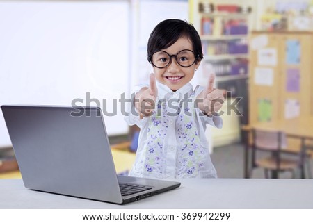 Lovely little girl with laptop computer on the table, showing thumbs up on the camera, shot in the classroom