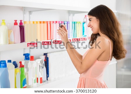 Side view of smiling female customer photographing cosmetic products on shelf in supermarket